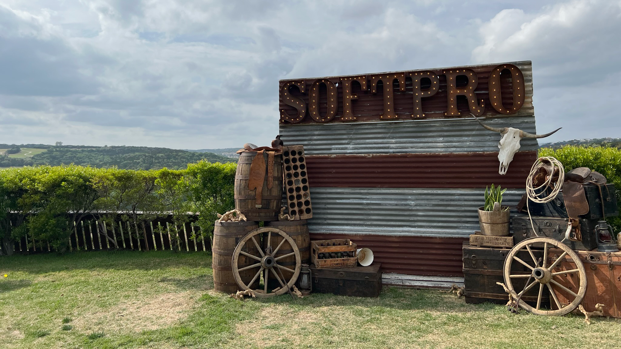 Rustic western-style display with a lit “SOFTPRO” marquee sign mounted on corrugated metal, surrounded by old wooden barrels, wagon wheels, crates, luggage, and a longhorn skull, set on grass overlooking rolling green hills under a cloudy sky.